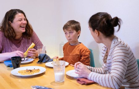 Familie aus zwei lesbischen Müttern und ihrem Sohn beim gemeinsamen Frühstück zu Hause, während der Junge Gesichter macht und lacht