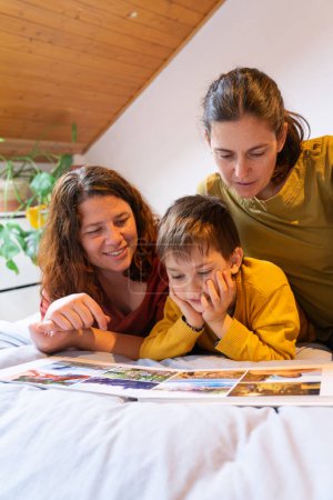 Familia LGBT de dos madres y un niño acostado en la cama mirando un álbum de fotos juntos