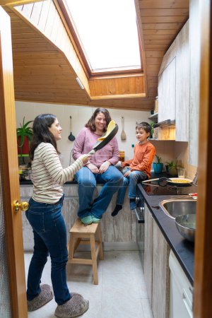 La famille LGBT cuisine des crêpes à la maison, retournant la crêpe dans la casserole. Deux mères et leur fils cuisinent ensemble