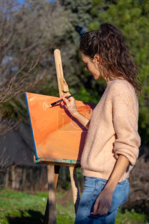 Woman brush painting an abstract painting outdoors with peach-colored acrylic paint