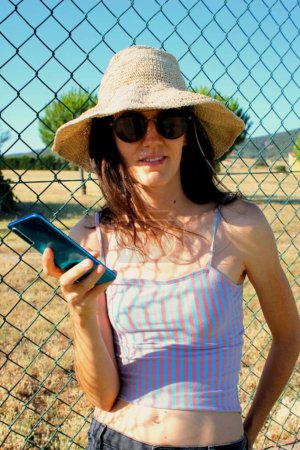 Mujer caucásica usando un sombrero de sol y gafas de sol y sosteniendo un teléfono móvil en su mano
