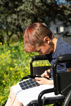 Caucásico niño de 8 años con discapacidad física en silla de ruedas usando un teléfono celular mientras se carga con un cargador solar en la naturaleza
