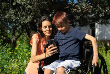 Mother teaching her son in a wheelchair something on the mobile in nature