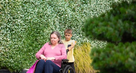 Boy carrying his grandmother in a wheelchair with a vertical garden background