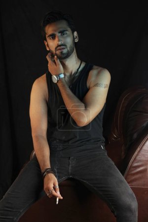 Foto de Hombre indio guapo con barba en jeans y camiseta negra - camisa con cigarro en la mano sentado en silla de cuero - Imagen libre de derechos