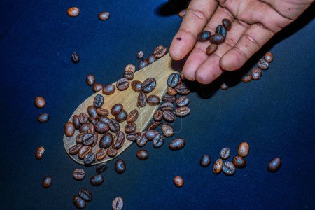 Black coffee beans are seen close up with a wooden spoon on a black cloth.