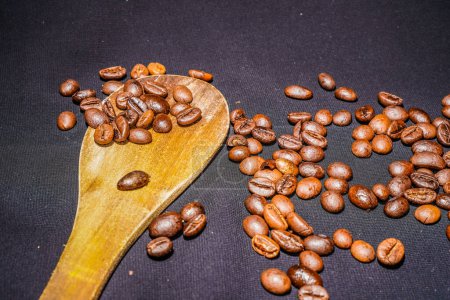 Black coffee beans are seen close up with a wooden spoon on a black cloth.