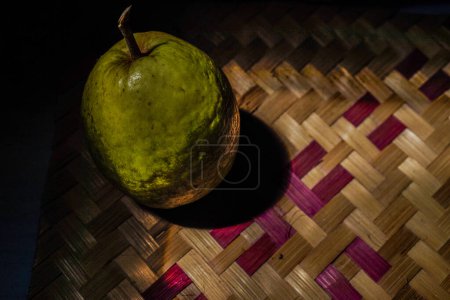 Foto de Fruto de guayaba aislado en bambú tejido con iluminación natural de la luz del sol, fondo oscuro para espacio de copia. - Imagen libre de derechos