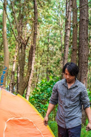 Javanese man is walking next to a tent set up in the middle of the forest