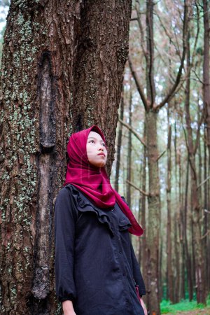 A Javanese woman wearing a headscarf is leaning against a tree in the forest with her gaze facing the sky.