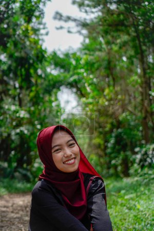 Asiática mujer en hijab es sentada en cuclillas en un camino rocoso en el bosque sonriendo a la cámara.