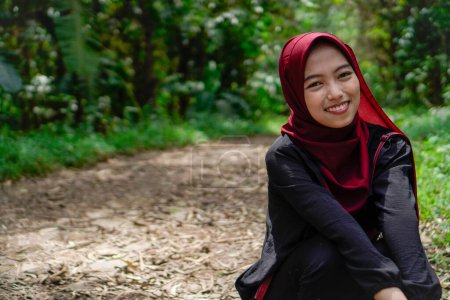 Asiática mujer en hijab es sentada en cuclillas en un camino rocoso en el bosque sonriendo a la cámara.