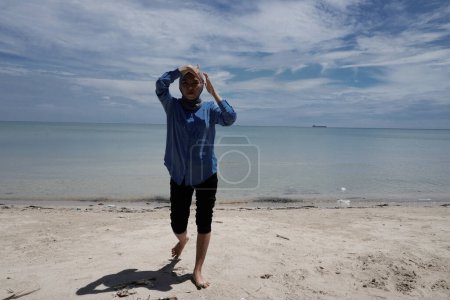 Asian woman in hijab is playing on the beach against a bright sky background with empty advertising space.