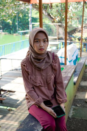 Javanese woman in hijab is sitting in the park with a smiling expression, summer vacation concept.