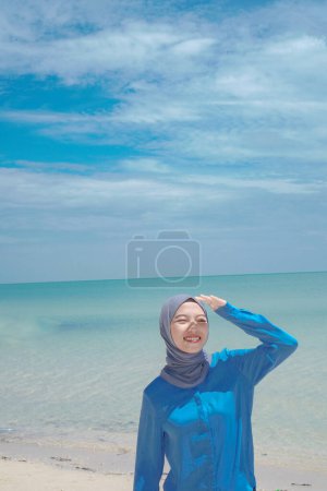 beautiful woman in hijab is standing with her hands covering her face from the hot sun tipi sea background of clear sky with empty space for advertising