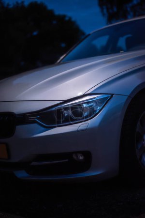Téléchargez les photos : Vue détaillée d'un phare BMW série 3 2012. Pris la nuit, un lampadaire met en valeur le capot et le phare du wagon argenté. - en image libre de droit