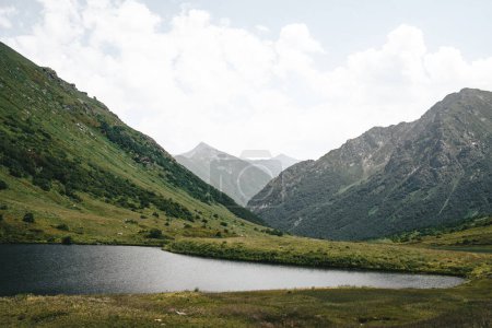 Foto de Pequeño lago de montaña entre picos verdes de montaña. Nubes, varias colinas, vista panorámica. Krasnaya Polyana, Montañas del Cáucaso. Hermoso paisaje con río y cordillera. - Imagen libre de derechos