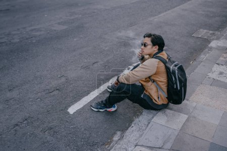 Photo for Young Asian Indonesian man wearing sunglasses sitting smoking on the highway sidewalk. Man addicted to cigarettes with copy space. Unhealthy background. - Royalty Free Image