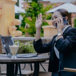 Beautiful positive Asian woman sits at an outdoor table of a cafe talking on the phone while waving her hand to call or greet someone. People and lifestyle concepts.
