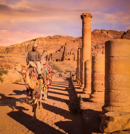Foto de Conductor de camello cabalgando por la calle Colonnaded, Petra, Jordania. Tumbas Reales en la distancia. - Imagen libre de derechos
