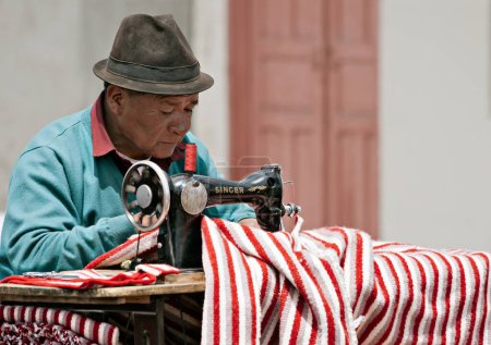 Foto de Un sastre callejero en el mercado de Saquisili, Ecuador, haciendo un poncho tradicional a rayas con su vieja máquina de coser Singer - Imagen libre de derechos
