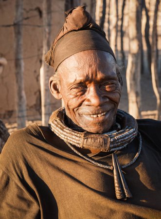 Foto de Himba anciano con traje tradicional y turbante sonríe frente a su cabaña de pueblo cerca de Opuwo, Región Kunene, Namibia. Al casarse, los hombres de Himba usan turbantes que nunca quitan.. - Imagen libre de derechos