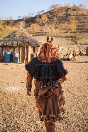 Foto de Mujer Himba en traje tradicional caminando hacia su cabaña en un pequeño pueblo ubicado cerca de Opuwo, Región Kunene, Namibia. Himba es una tribu tradicional en África. - Imagen libre de derechos