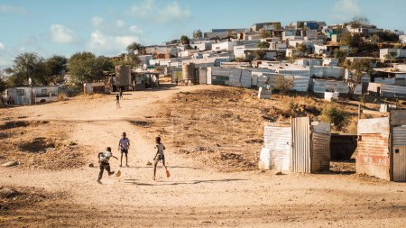 Téléchargez les photos : Groupe de garçons jouant au football parmi les huttes rugueuses de Katutura, Windhoek, région de Komas, Namibie. Katutura est un canton créé dans les années 1950 pour séparer la population noire de Windhoek. - en image libre de droit