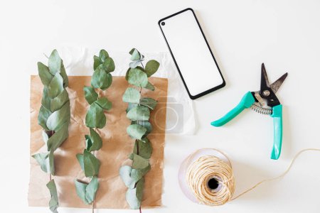 Téléchargez les photos : Espace de travail fleuriste minimal avec téléphone portable, papier artisanal et branches d'eucalyptus sur fond blanc - en image libre de droit