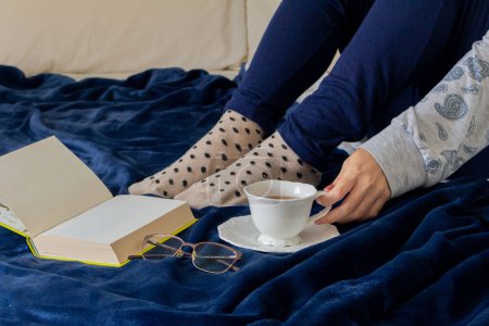 Téléchargez les photos : Jeune femme en pyjama, boire du thé et lire au lit - en image libre de droit