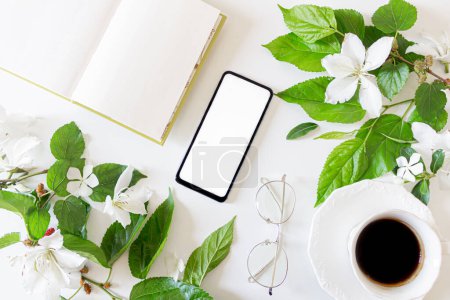 Photo for Home office desk with blank screen mobile phone with copy space. White flowers and blackberry tree leaves frame. Glasses, cup of coffee and open book on white background. Flat lay. Spring composition. - Royalty Free Image