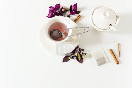 Foto de Copa de composición de té de jengibre con hojas púrpura y rosa, canela, colador de té, bolsa de té sobre fondo blanco. - Imagen libre de derechos