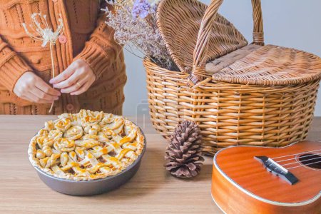 Foto de Composición otoñal con flores secas en cesta de picnic, ukelele, tarta de manzana decorada y mujer joven de pie sobre el fondo - Imagen libre de derechos