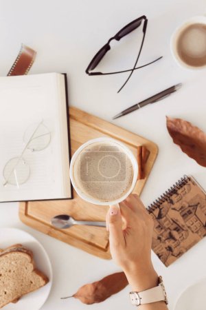 Foto de Mano femenina sosteniendo taza de café. Otoño Composición del desayuno con libro, anteojos y hojas secas alrededor. - Imagen libre de derechos