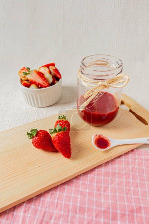 Téléchargez les photos : Marmelade de fraises sur un pot, une petite cuillère blanche avec un peu de confiture, et des fraises fraîches sur un bol. Fond clair. - en image libre de droit