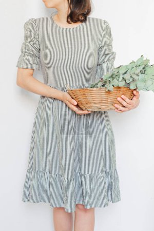 Photo for Young pretty woman in striped vintage dress holding a straw basket with an eucalyptus bouquet on white background. Florist minimal concept. - Royalty Free Image
