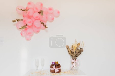 Photo for Romantic concept. Chocolate cake with two empty glasses, bouquet of dried flowers and pile of pink balloons - Royalty Free Image