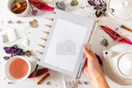 Photo for Tea time feminine frame composition with female hand holding a planner, tea pot, cinnamon, cup, tea strainer, purple and pink leaves and dried plants on white background. Flat lay, top view. - Royalty Free Image