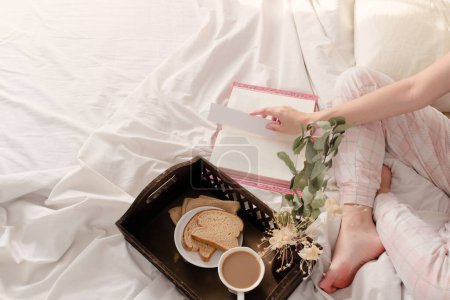 Téléchargez les photos : Femme lisant un livre et petit déjeuner sur plateau vintage en bois foncé au lit avec drap beige clair et oreillers. - en image libre de droit