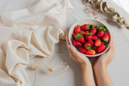 Photo for Young female hands holding a bowl full of strawberries on a styled light background. - Royalty Free Image