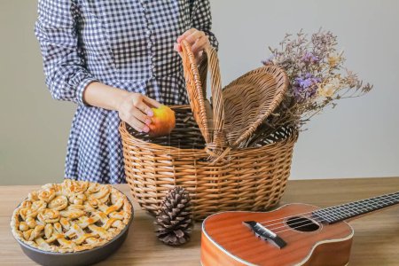 Foto de Foto recortada de una mujer joven poniendo manzana en la cesta de picnic. Tarta de manzana decorada con ukelele y cono de pino sobre mesa de madera - Imagen libre de derechos