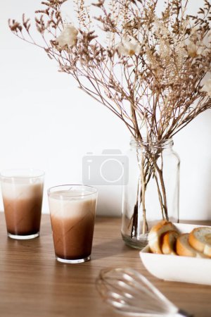 Foto de Café Dalgona con leche, ramo de flores secas en jarrón y tostadas en plato sobre mesa de madera. - Imagen libre de derechos