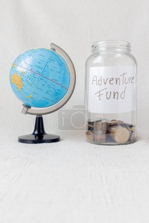 Foto de Tarro de vidrio con el mensaje: "fondo de aventura" y algunas monedas dentro junto a un globo terráqueo sobre fondo blanco. Concepto de viaje. - Imagen libre de derechos