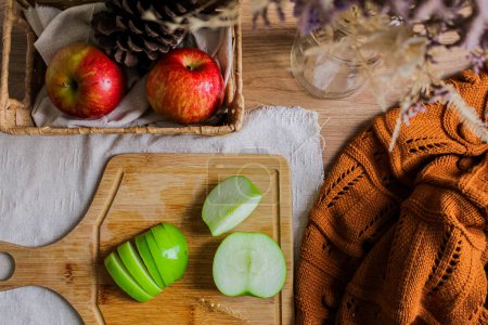 Foto de Composición otoñal con manzanas verdes en rodajas sobre tabla de madera, cesta de paja con manzanas rojas y cono de pino y suéter marrón - Imagen libre de derechos