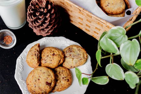 Photo for Cookies on a plate, pine, glass of milk, straw basket, plant leaves on black background. - Royalty Free Image