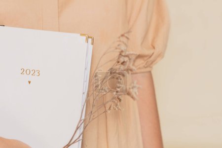 Photo for Cropped shot of woman wearing a beige dress, holding a 2023 planner and wildflowers. Aesthetic composition. - Royalty Free Image