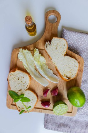 Foto de Cocina Mediterránea - Tabla de cortar preparada para hacer una buena ensalada seguida de pan como plato de inicio. Aceite de oliva, acelga, albahaca, ajo, limón y un trozo de pan fresco. - Imagen libre de derechos