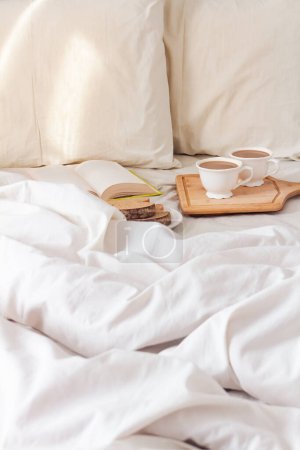 Photo for Morning romantic breakfast concept. Fresh sliced bread with cups of coffee with book in bed with light beige sheet and pillows. - Royalty Free Image