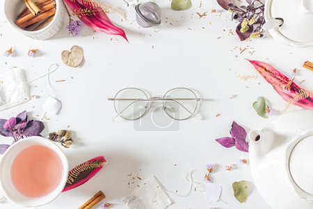 Photo for Tea time frame composition with eyeglasses in the middle of frame with teapot, cinnamon, cup, tea strainer, purple and pink leaves and dried plants on white background. Flat lay, top view. - Royalty Free Image