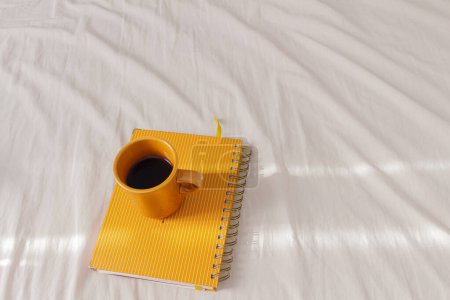 Photo for Top view of coffee cup on the top of yellow planner on bed sheet - Royalty Free Image